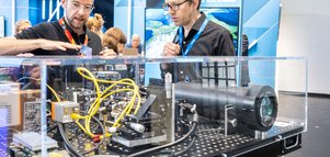 Two men look at complex machines with cables and components in a display case at a trade fair stand and chat.