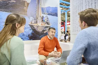 Sales talk at a trade fair stand on the subject of sailing trips. A couple is being advised by a man with a laptop in front of him. In the background is a poster with a sailing boat.