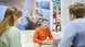 Sales talk at a trade fair stand on the subject of sailing trips. A couple is being advised by a man with a laptop in front of him. In the background is a poster with a sailing boat.