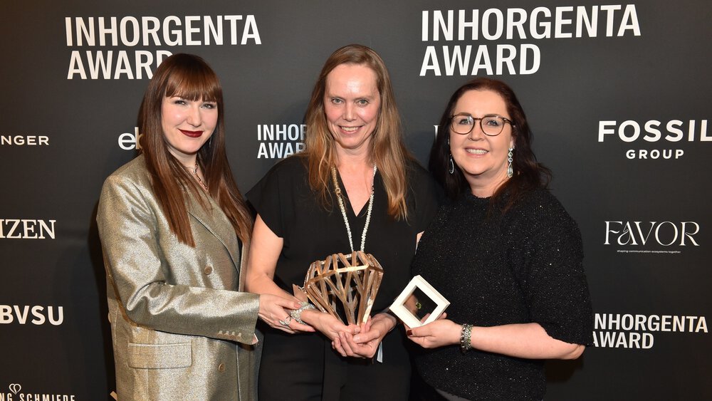 Three smiling women hold the trophy for the “Designer of the Year” category at the INHORGENTA AWARD Gala in front of a logo wall.