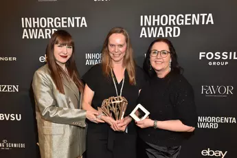 Drei lächelnde Frauen halten die Trophäe der Kategorie „Designer of the Year“  bei der INHORGENTA AWARD-Gala vor einer Logowand.