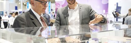 Zwei Männer in Anzügen besprechen Produkte, die in einer Glasvitrine an einem Messestand ausgestellt sind.
