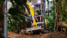 A man operates a mini excavator in a green garden landscape with many plants and trees.