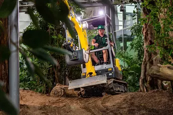 Ein Mensch bedient einen Minibagger in einer grünen Gartenlandschaft mit vielen Pflanzen und Bäumen.