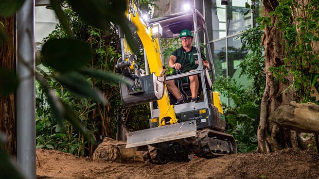 Ein Mensch bedient einen Minibagger in einer grünen Gartenlandschaft mit vielen Pflanzen und Bäumen.