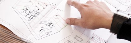 A hand points to construction plans lying on a table with sketches of furniture and the layout of a trade fair stand.