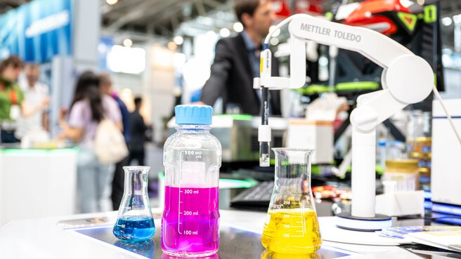 Three transparent containers stand next to each other. The one on the left contains a blue liquid, the one in the middle a pink liquid and the one on the right a yellow liquid