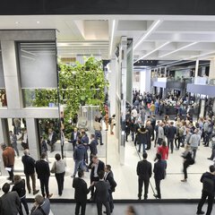 Blick über einen gut besuchten Aufenthaltsbereich für Messebesucher der BAU mit Sitzmöglichkeiten und Pflanzen, daneben ein Durchgang zu weiteren Messebereichen.