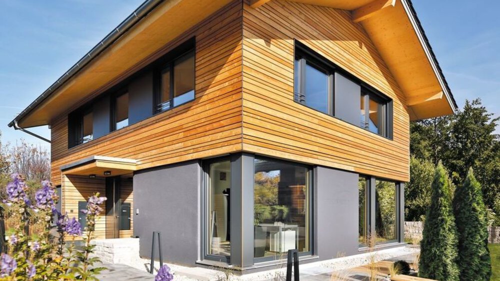Modernes zweistöckiges Haus mit Holz- und Graufassade, großen Fenstern und umgebendem Grün unter einem klaren blauen Himmel.