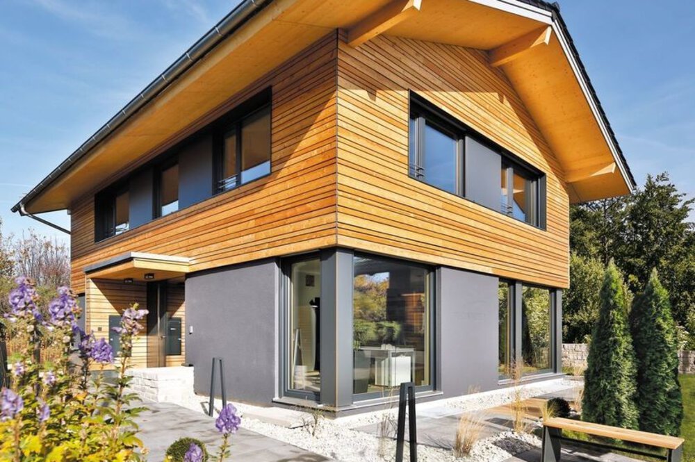 Modernes zweistöckiges Haus mit Holz- und Graufassade, großen Fenstern und umgebendem Grün unter einem klaren blauen Himmel.