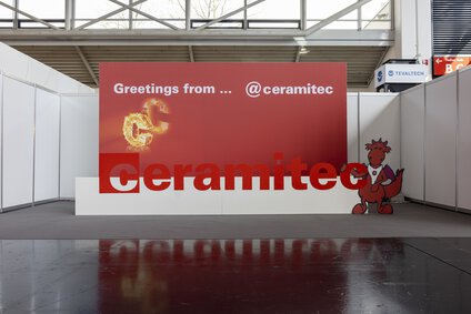 auf dem Bild erkennt man eine große rote Wand auf der ceramitec steht, vor der Wand ist ein Drache aus Pappe der den roten Schriftzug ceramitec hochhält