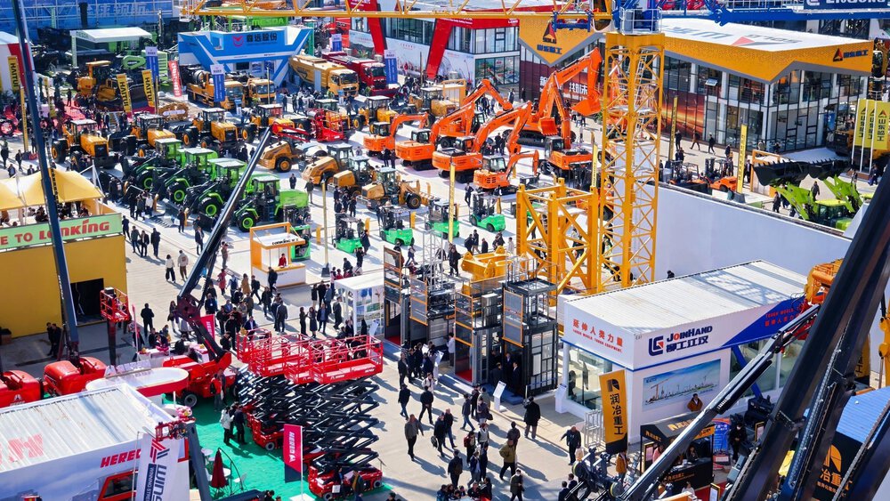 A bustling outdoor trade show with crowds of people, tractors, construction equipment, and colorful company booths.