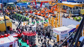 A bustling outdoor trade show with crowds of people, tractors, construction equipment, and colorful company booths.