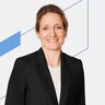 Frau mit hellbraunem Haar in einem schwarzen Blazer, lächelnd vor einem weißen Hintergrund mit blauen Linien.