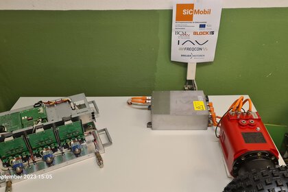 Auf einem Tisch angeordnete elektronische Komponenten und Geräte vor einer grünen Tapete. An der Wand hängt ein Zettel mit der Aufschrift 