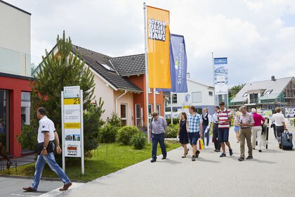 Menschen gehen an verschiedenen Musterhäusern vorbei und halten Flyer und Broschüren mit Informationen zum Thema Wohnen in der Hand.