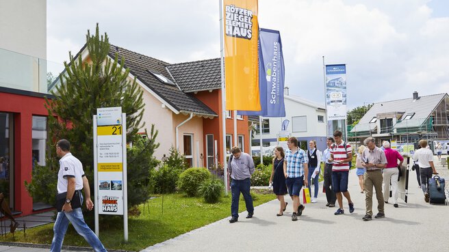 Menschen gehen an verschiedenen Musterhäusern vorbei und halten Flyer und Broschüren mit Informationen zum Thema Wohnen in der Hand.