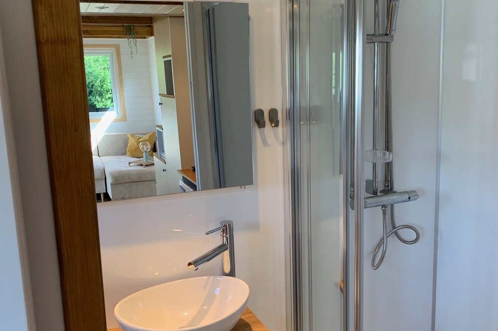 Ein Badezimmer mit Waschbecken und Dusche, neben einem Zimmer mit Bett und einem Fenster mit Blick ins Grüne.
