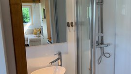 Ein Badezimmer mit Waschbecken und Dusche, neben einem Zimmer mit Bett und einem Fenster mit Blick ins Grüne.