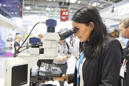 A woman in a black blazer looks through a microscope at a trade fair stand. In the background, the A3 exhibition hall can be seen out of focus.