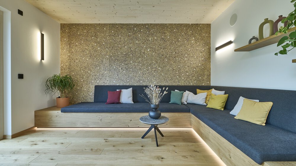 Elegant schlichtes Wohnzimmer mit grauem Ecksofa, bunten Kissen, Couchtisch aus Holz, Pflanzen und strukturierter Wand.