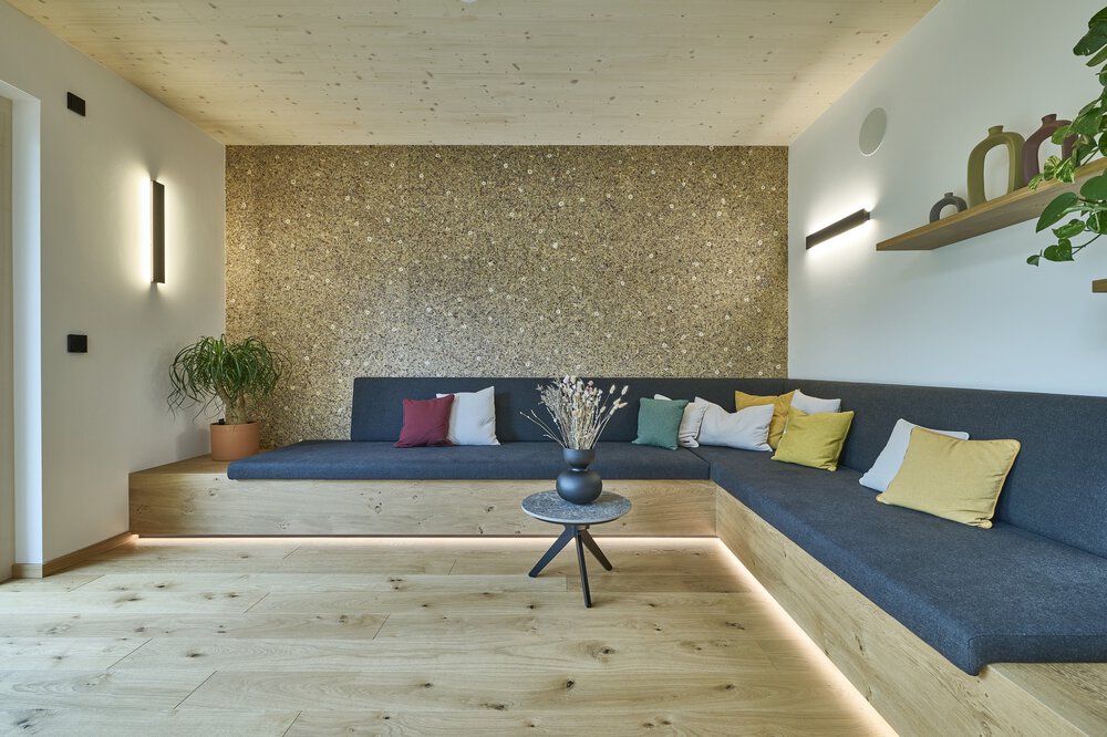 Elegant schlichtes Wohnzimmer mit grauem Ecksofa, bunten Kissen, Couchtisch aus Holz, Pflanzen und strukturierter Wand.