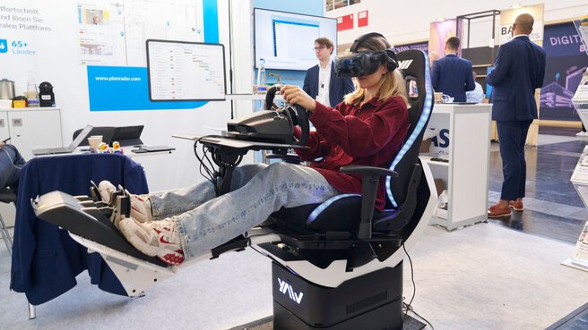 A woman with VR glasses on her head sits in a chair with a steering wheel and foot pedals.