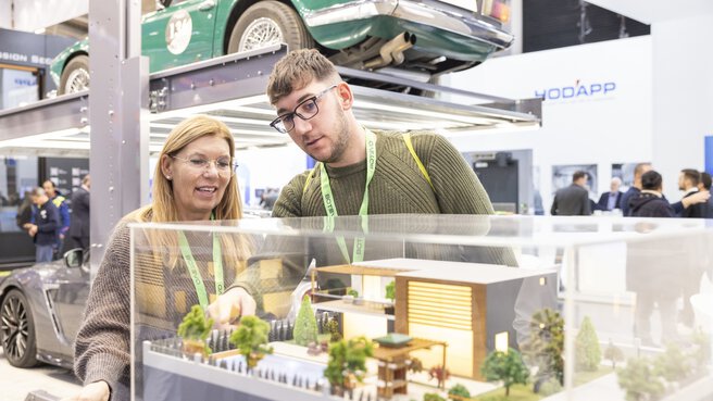 A woman and a man are looking at a display case with a model house. Two cars are on display behind them.
