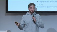 Ein Mann im grauen Hoodie spricht in ein Mikrofon und hält dabei einen Clicker. Hinter ihm ist eine Präsentationsfolie zu sehen.