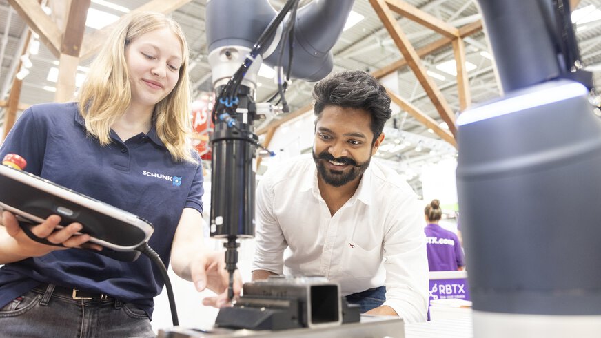 An exhibitor demonstrates a robot arm to a visitor at an exhibition stand. The visitor watches in fascination.