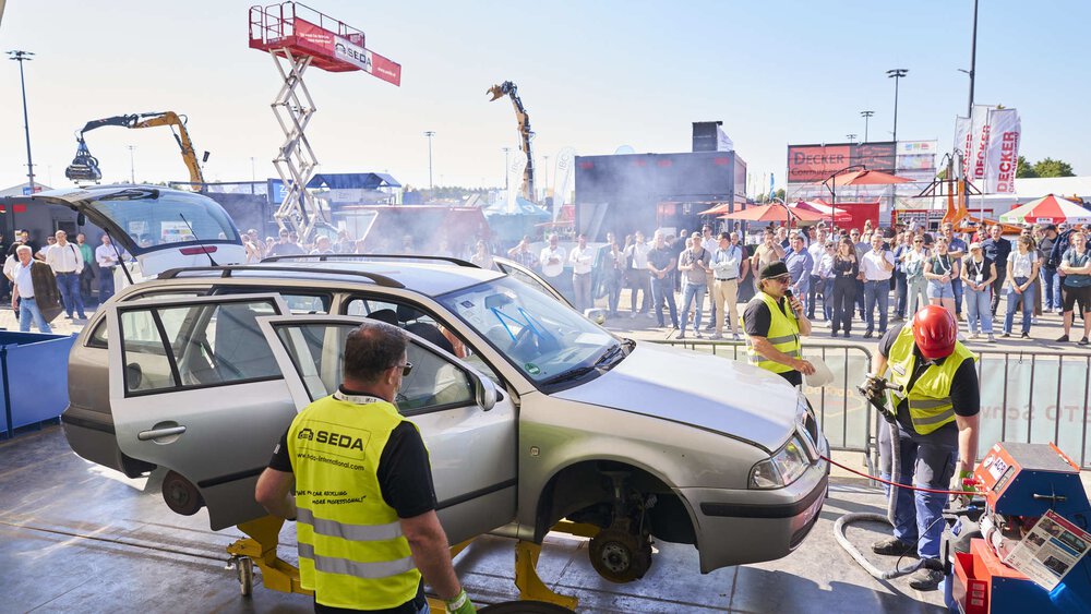 Demonstration im Freigelände zum Thema Fahrzeugverwertung. Arbeiter mit gelben Westen stehen um ein aufgebocktes Auto mit demontierten Rädern.