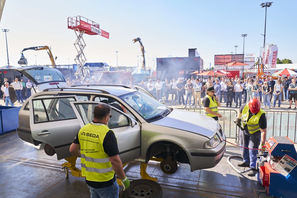 Demonstration in the outdoor area on the subject of vehicle recycling. Workers with yellow vests stand around a jacked-up car with dismantled wheels.