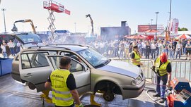 Demonstration in the outdoor area on the subject of vehicle recycling. Workers with yellow vests stand around a jacked-up car with dismantled wheels.