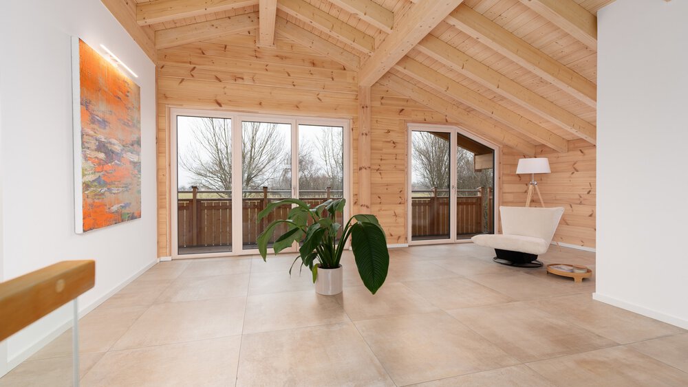 Ein geräumiger Raum mit Holzdecke und -wänden mit großen Fenstern zum Balkon, einer Topfpflanze, einem modernen weißen Sessel und einem farbenfrohen Gemälde an der Wand.