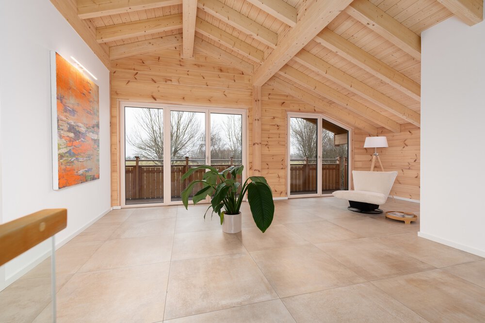 Ein geräumiger Raum mit Holzdecke und -wänden mit großen Fenstern zum Balkon, einer Topfpflanze, einem modernen weißen Sessel und einem farbenfrohen Gemälde an der Wand.