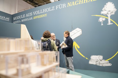 Mehrere Personen unterhalten sich an einem Stand auf der BAU. Im Hintergrund wirbt der Aussteller für Module aus Vollholz für mehr Nachhaltigkeit.