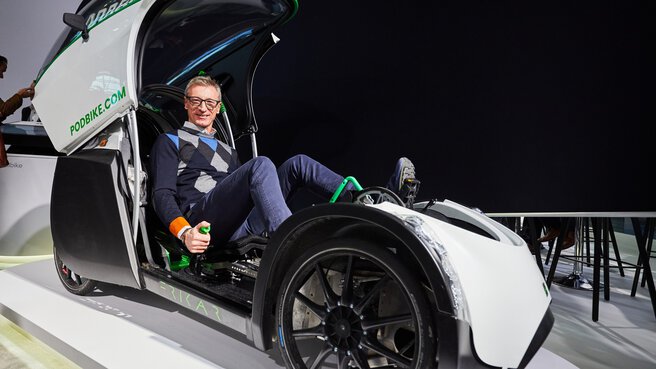 Ein Mann sitzt lächelnd in einem futuristischen, elektrischen Liegefahrrad, dass an einem Messestand ausgestellt ist.