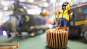 A toy figure wearing a helmet and safety vest stands on a toroidal choke and holds on to the wire. In the background, a model helicopter can be seen out of focus.