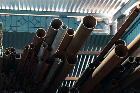 Stacked metal pipes and profiles in various diameters and shapes, stored under a corrugated metal roof inside a hall.