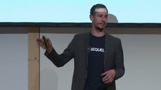 Eine Person im Jackett und T-Shirt spricht auf der Bühne und hält ein Mikrofon sowie einen Clicker.