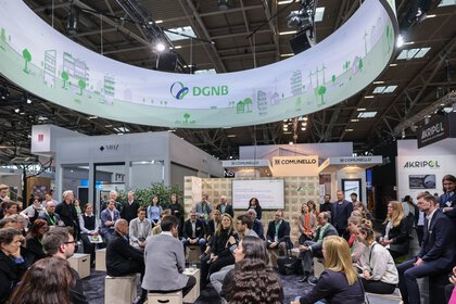 Zahlreiche Besucher der BAU sitzen und stehen in einem Kreis unter einem runden Werbebanner der „DGNB“ und lauschen einer Diskussion in der Mitte.