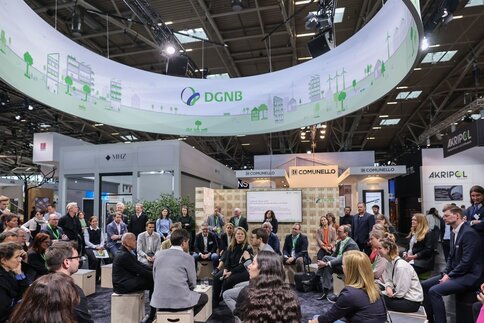 Zahlreiche Besucher der BAU sitzen zu einer Diskussionsrunde auf Holzkisten im Kreis. Über ihnen ist ein runder Werbebanner der 