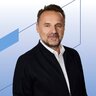 Ein Mann mittleren Alters mit Bart, der eine dunkle Jacke und ein weißes Hemd trägt, vor einem blauen geometrischen Hintergrund.