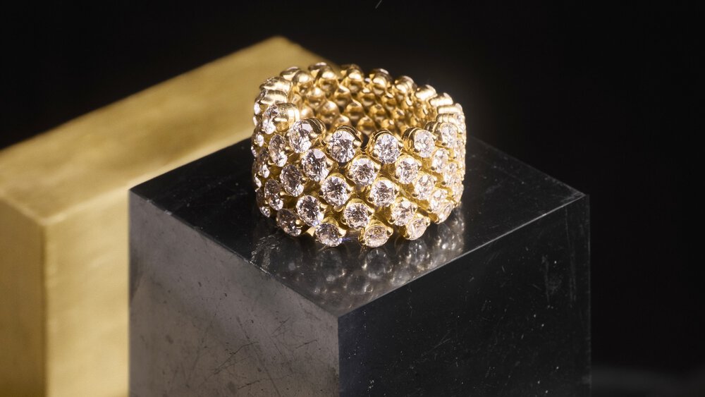 A gold ring set with several rows of diamonds sits on a black base against a dark background.