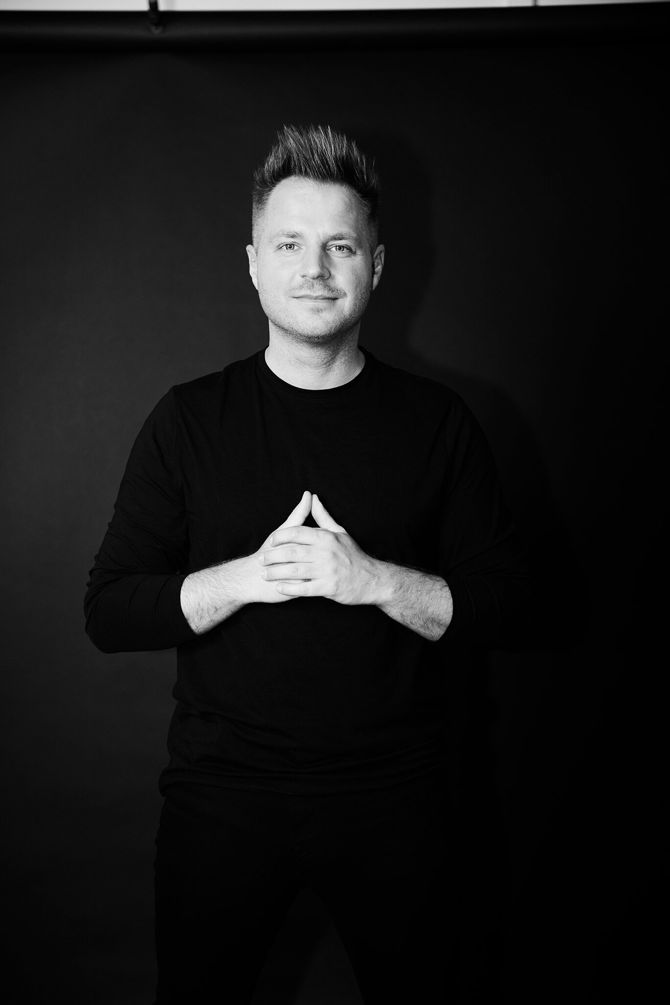 A smiling man in a black long-sleeved shirt stands with his hands folded in front of a dark background and looks ahead.