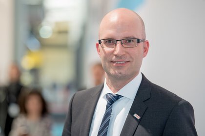 A man in a suit with a tie and glasses stands smiling in an interior space against a blurred background.