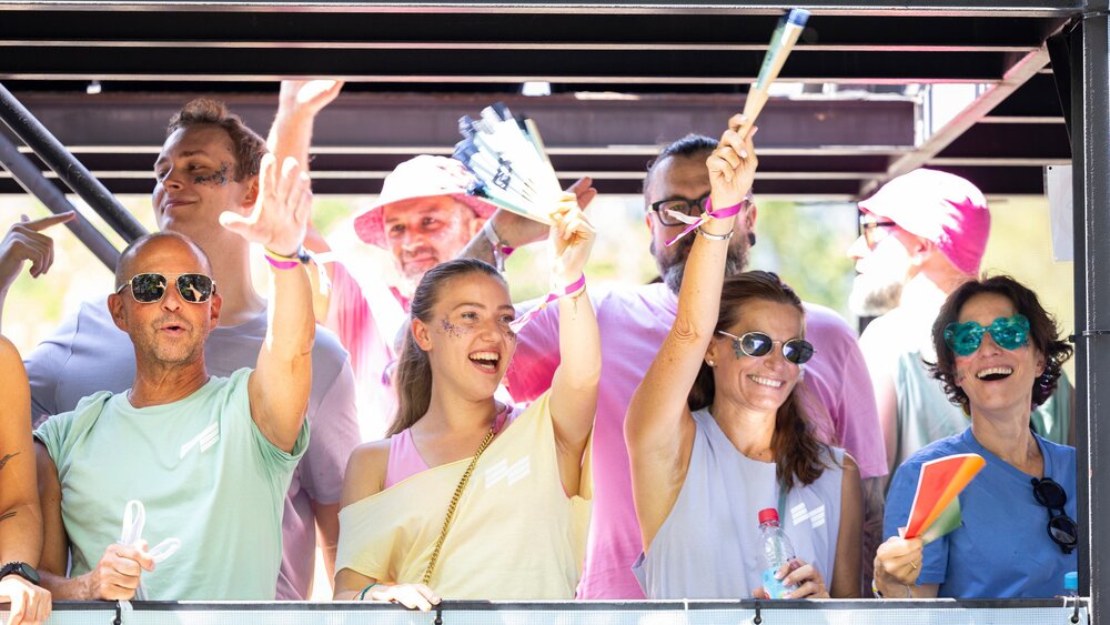 A group of people smiling, waving, and cheering at an outdoor event in bright sunshine.