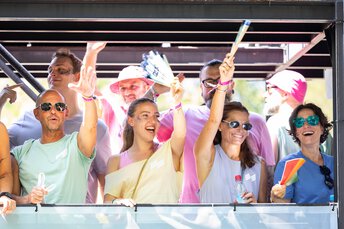 A group of people smiling, waving, and cheering at an outdoor event in bright sunshine.