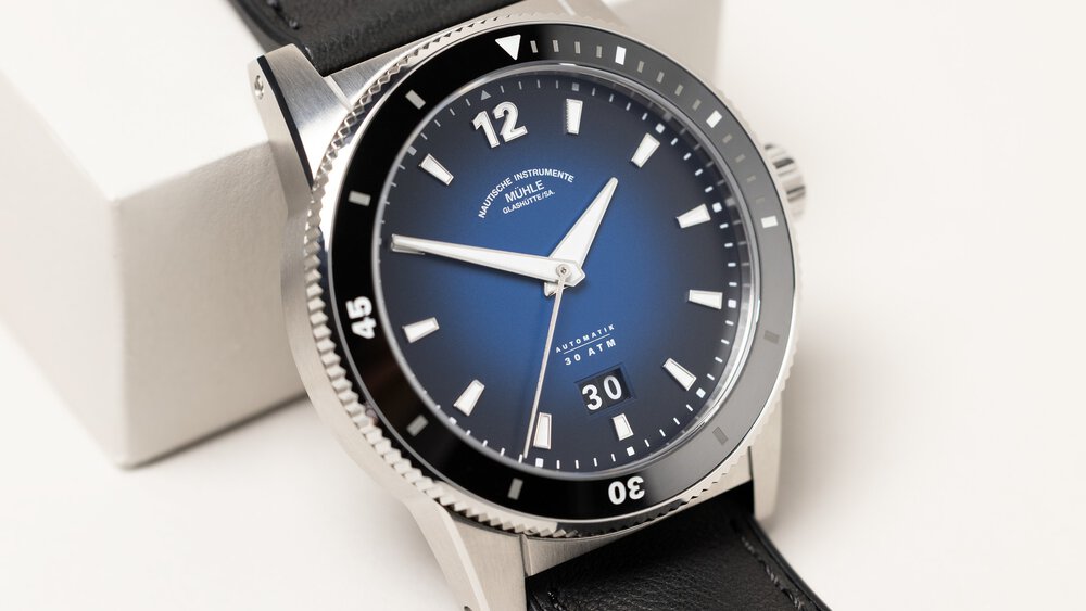 Close-up view of a delicate wristwatch with a dark blue shaded dial with day display, a jagged bezel, and black straps.