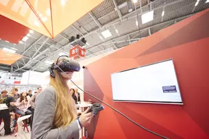 A woman with long hair wears virtual reality goggles and holds controllers. She is standing in front of an empty monitor on a red wall at an exhibition stand.